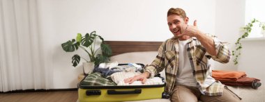 Yakışıklı, karizmatik bir gencin portresi, yüzünün yanında telefon işareti, bavuldaki giysilerini gösteriyor, tatile çıkıyor, birlikte tatile çıkıyor, seyahat acentesinin reklamını yapıyor..