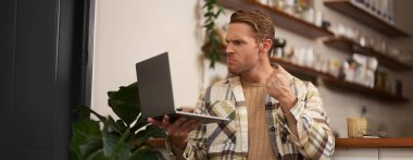 Kafedeki kızgın adamın portresi, dizüstü bilgisayar, ekrana bakıyor ve görüntülü konuşma sırasında yumruğunu gösteriyor, online toplantı..