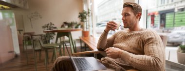 Çalışan insanlar ve kamusal alan kavramı. Genç yakışıklı adam, ux ui tasarımcı, laptopuyla kafede oturuyor, cappuccino içiyor, ciddi bir yüzle ekrana bakıyor..