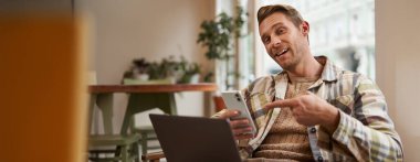 Kafedeki heyecanlı adam, elinde telefonla ekranı göstererek, bir uygulama öneriyor, birlikte çalışma alanında mobil cihaz ve dizüstü bilgisayar kullanıyor. Serbest çalışma alanı ve uzak çalışma alanı kavramı.