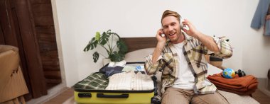 Genç mutlu bir adamın portresi, kulaklıkla müzik dinliyor, otel odasında bavuluyla oturuyor, tatilde giysilerini açıyor, turist tatile çıkıyor. Yaşam tarzı ve insan kavramı