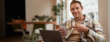 Serbest çalışan genç bir adamın portresi, kafede çalışıyor, dizüstü bilgisayarla sandalyede oturuyor, telefon ekranını işaret ediyor, telefonundaki bir şeyle ilgileniyor..