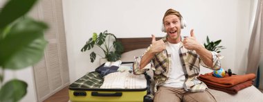 Mutlu, tatmin olmuş genç bir adam, onaylama jesti yapıyor, kablosuz kulaklıklarla oturuyor, bavullarını paketliyor, tatilde bavuluyla otelde turist. Yaşam tarzı kavramı