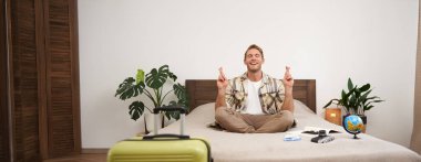 Küre ve kameralı genç adam, yatakta oturup meditasyon yapıyor, bavulunu hazırlıyor, yanında duruyor, tatile çıkıyor, tatilde iyi hava için dua ediyor, parmaklarını çaprazlayarak dilek diliyor..