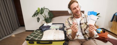 Mutlu turist, genç adam pasaport ve bilet gösteriyor, tatile gidiyor, valiziyle yatağa oturuyor, yaz tatili planları yapıyor..