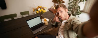 Yakışıklı genç adam çalışırken ya da laptopla uzaktan çalışırken selfie çekiyor, telefon işaretini gösteriyor ve cep telefonu kamerasına gülümsüyor..