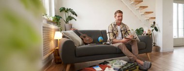 Neşeli yakışıklı adam tatil için hazırlanıyor, tatile çıkıyor, yazıyor, not alıyor, seyahat için eşyaların listesini yapıyor, bavulunu kıyafetlerle dolduruyor..