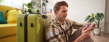 Bavullu bir adam portresi, yere oturmuş, cep telefonuyla oynuyor, ciddi görünüyor. Turizm ve halk konsepti