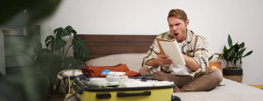 Kızgın bir adamın portresi, bavul hazırlıyor, tatil için kontrol listesiyle defterde hayal kırıklığına uğramış görünüyor, tatile çıkmaya hazırlanıyor. Turist ve insan konsepti