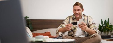 Biletleri ayırtan genç adamın portresi, tatile gitmek, kredi kartı ve akıllı telefon tutmak, giysilerle dolu çantanın yanında oturmak, tatile çıkmaya hazır olmak..