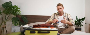 Yakışıklı bir adamın portresi, turist, tatil biletlerine bakıyor seyahat acentesinde yer ayırtıyor, yatakta oturuyor ve giysilerini bavuluna koyuyor..