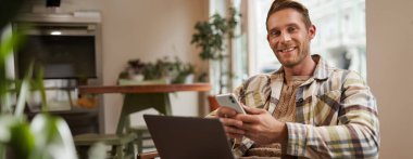 Genç, yakışıklı bir adamın portresi, kafeterya ziyaretçisi, dizüstü bilgisayarlı ve akıllı telefonlu, kameraya gülümseyen, uzaktan çalışan serbest çalışan biri..