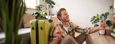 Gülümseyen yakışıklı bir adamın portresi, barış işareti gösteriyor, akıllı telefondan selfie çekiyor, bavulla oturuyor, tatile çıkıyor. Turizm ve seyahat acentası kavramı