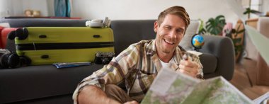 Seyahat haritalı mutlu genç adam, seyahatini planlıyor, turneye çıkıyor, bavuluyla evde oturuyor..