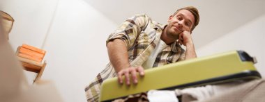 Çantayı kapatmaya çalışan komik bir adam resmi. Elbise yığını, bavul, eşyaları çantaya koyuyor. Turizm ve gezgin konsepti