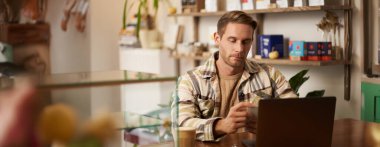 Kafe sahibi portresi, kafeteryada dizüstü bilgisayarla oturan genç bir adam, veri projesi üzerinde çalışıyor, ekrana odaklı bakıyor. Yaşam tarzı ve iş konsepti