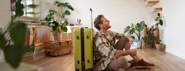 Gülümseyen genç adamın portresi, uçak biletleri, tatile gitmek, oturma odasında yeşil bavulla oturmak..