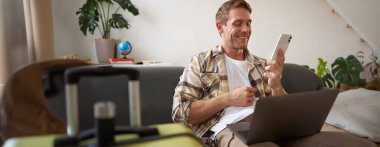 Gülümseyen yakışıklı bir adam tatile çıkıyor, telefonla konuşuyor, dizüstü bilgisayarını tutuyor, yurt dışına seyahat için bavul hazırlıyor..