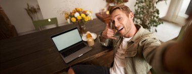 Yakışıklı adam cep telefonunda dizüstü bilgisayarla selfie çekiyor, çağrı el işareti yapıyor, kameraya gülümsüyor, sosyal medya uygulamasına fotoğraf yüklüyor, kafede oturuyor..