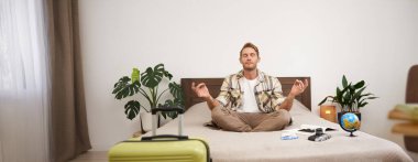 Genç, rahat bir turist portresi, bacak bacak üstüne atıp meditasyon yapan bir adam, bavulunu, küreyi ve kamerasını topladı, tatile çıkıyor, seyahate çıkmadan önce sakinleşiyor..