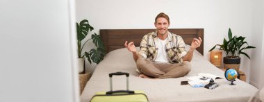 Meditasyon yapan genç bir adamın portresi, bavul hazırlamak, dünya, kamera ve uçak biletleriyle yatakta oturmak, yurt dışına çıkmadan önce dinlenmek. Turizm konsepti