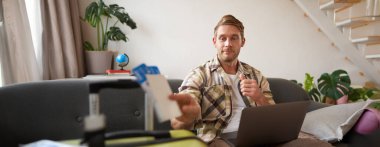 Mutlu genç adamın portresi iki kişilik tatil rezervasyonu, uçak biletleri, dizüstü bilgisayara bakması, oturma odasında bilgisayarla oturması. Seyahat ve turizm konsepti