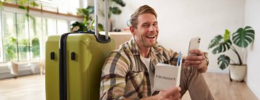 Gülümseyen yakışıklı turistin portresi, elinde pasaport ve uçak bileti olan bir adam, bavulla oturuyor. Turizm ve seyahat kavramı