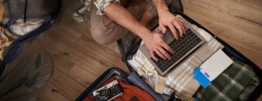 Erkek eli yakın çekim, dizüstü bilgisayar kullanan turist, açık bavulla yerde oturuyor, tatil için bavullarını hazırlıyor, iki uçuş bileti, kamera ve giysi var..