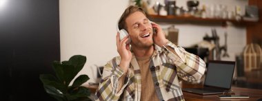 Yakışıklı, gülümseyen genç bir adam, dijital göçebe, açık alanda birlikte çalışıyor, kafeteryada çalışıyor, kablosuz kulaklıkla müzik dinliyor ve şarkı söylüyor..