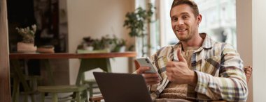 Gülümseyen, kahve dükkanında rahatlayan yakışıklı genç bir adamın portresi, kafeteryada ortak çalışma alanından uzaktan çalışma, dizüstü bilgisayar ve akıllı telefon kullanma, kameraya bakma ve tatmin olmuş yüzle baş parmaklarını gösterme.