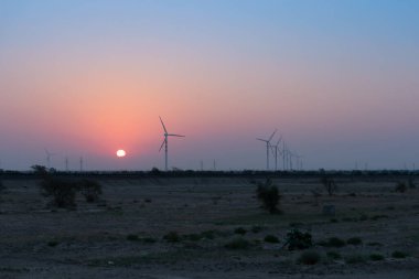 Çöl semalarında şafak vakti rüzgar değirmenleri oluşturarak yerel insanlar tarafından tüketilmek üzere alternatif çevre dostu yeşil enerji üretiyorlar. Güneş Thar Çölü, Rajasthan, Hindistan 'da doğuyor..