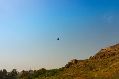 Zipline tourist flying over Rao Jodha Desert Rock Park, Jodhpur, Rajasthan, India. Near the historic Mehrangarh Fort , park contains ecologically restored desert and land vegetation, a tourist spot. clipart