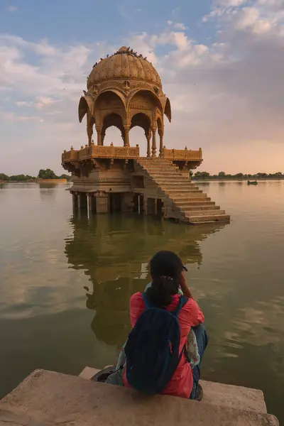 stock image Indian female traveller, woman photographer taking picture of Chhatris and shrines with reflection of them on the water of Gadisar lake. Sun set and colorful clouds on sky. Jaisalmer, Rajasthan,India.