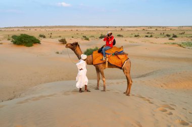 Female tourist taking picture while riding camel, Camelus dromedarius, at sand dunes of Thar desert, Rajasthan, India. Camel riding is a favourite activity amongst all tourists visiting here.