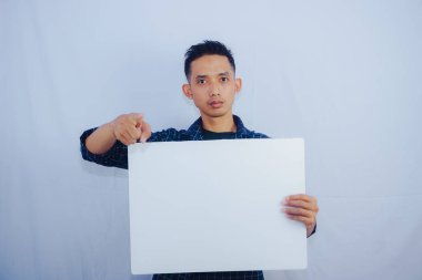 Reklam Kavramı. Beyaz stüdyo arka planında boş tahta tutan Asyalı adamın portresi. İlan için beyaz kare kağıt pankartlı bir adam duruyor, kameraya gösteriyor.