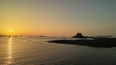 Saint-Malo, Brittany, Fransa yakınlarındaki sakin deniz üzerinde gün batımı manzarası, suların üzerinde süzülen siluetli yelkenliler ve ufukta antik bir kıyı kalesi. Huzurlu deniz manzarası