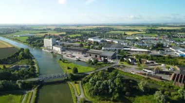 Yakınındaki siloları, tarlaları ve yaz mevsiminde gökyüzünün altında hafif sanayi bölgesiyle huzurlu bir tarım arazisi boyunca süzülen bir kanal görüntüsü. Fransa 'da Aire-sur-la-Lys yakınlarında çekildi..