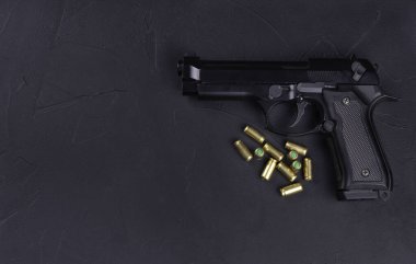 Siyah yarı otomatik bir tabancanın yakın plan görüntüsü ve koyu dokulu bir yüzeye yerleştirilmiş birkaç pirinç mermi. Tabanca bir açıyla yerleştirilmiş, kabzasını ve tetiğini vurguluyor, cephane de yakınlarda dağılıyor. Bu fotoğraf için ideal.