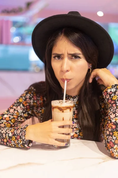 angry looking at the camera and having a drink with a straw, facial expression and drinks, young latin woman with long hair and fashionable hat, beauty and lifestyle