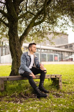 lifestyle of young latin man in a park in the shade of a tree wears a casual jean jacket, attractive and cheerful male model, sunny day