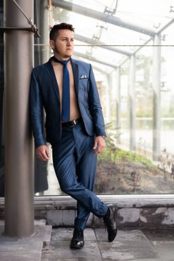 Kısa saçlı poz veren genç adam mavi bir takım elbise, kravat ve güneş gözlüğü takıyor, yaşam tarzı ve iş modası, şehir manzarası