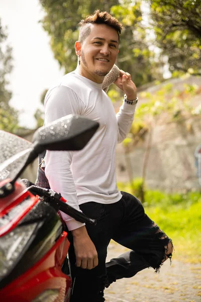 details of young latin man with short hair and casual clothes standing near modern motorcycle, transportation and adventure on the road, glamorous male model