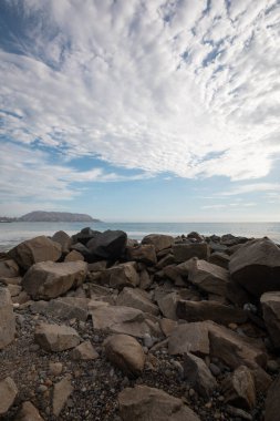 Gökyüzünde bulutlar, sahilde kayalar, gündüz turistik yerler, doğayla iç içe olan manzara, mekanın güzelliği.