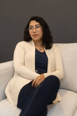 Bir kanepede dinlenen kısa saçlı güzel Latin bir kadın günlük kıyafetler ve gözlük takıyor, profesyonel ve manken yaşam tarzı, moda