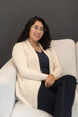 Kanepede oturan kısa saçlı genç bir Latin kadın günlük kıyafetler ve gözlük takıyor. Profesyonel bir model gibi gülümsüyor.