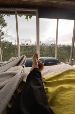 Rahat ve rahat bir yaşam tarzı, battaniyeli bir yatak odası ve arka planda bitkiler olan büyük pencereli, çıplak ayaklı bir insan yatağı.