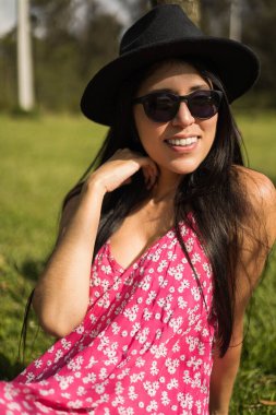 Doğanın tadını çıkaran göz kamaştırıcı genç Latin kadın, kadın desenli elbise, güneş gözlüğü ve şapka giyen manken, güzellik ve moda.