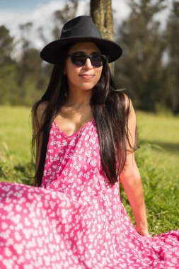 Güneşli bir gün, çimenlerde oturmuş doğanın tadını çıkaran genç bir Latin kadın, kadın elbisesi, güneş gözlüğü ve şapka takan bir manken, güzellik ve rahat moda.