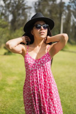Güneşli bir yaz günü, parkın doğası gereği genç Latin bir kadın yüzü, kadın elbisesi, güneş gözlüğü ve şapka takan bir model, yaşam tarzı ve güzellik.