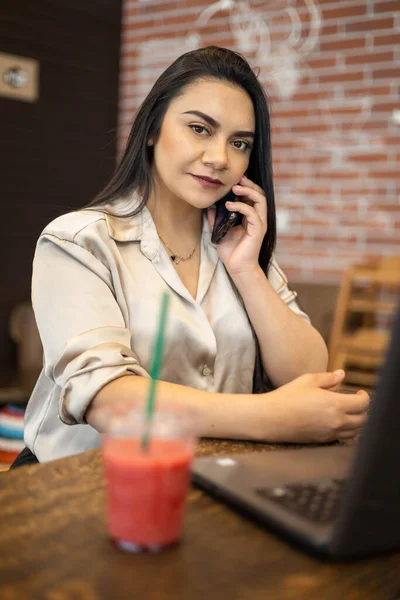ラテン語の忙しい若い女性が正式な着用と化粧をしてドリンクを飲みながら座って電話で携帯電話を使ってラップトップ テクノロジー ビジネスの隣で — ストック写真